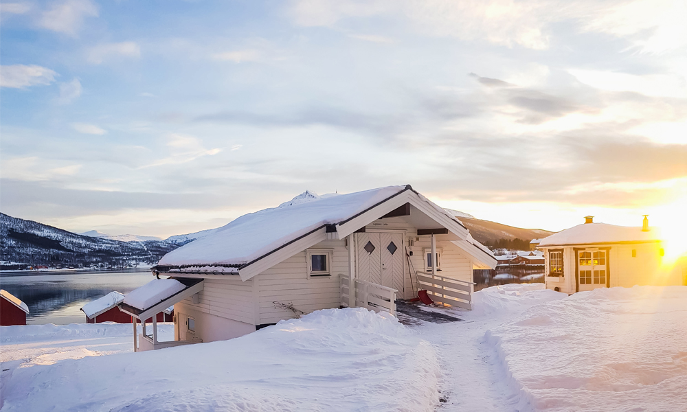 alt=“White small house and small sauna building surrounded by snow in Gibostad town, Northern Norway”