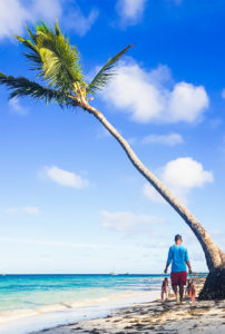 alt="man walking along the beach carrying red fish and passing a tall palm tree"
