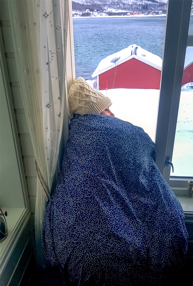 alt=“Paper Pelicans girl wrapped up in duvet and hat looking outside the window at snow and red wooden houses in Gibostad town, Northern Norway”