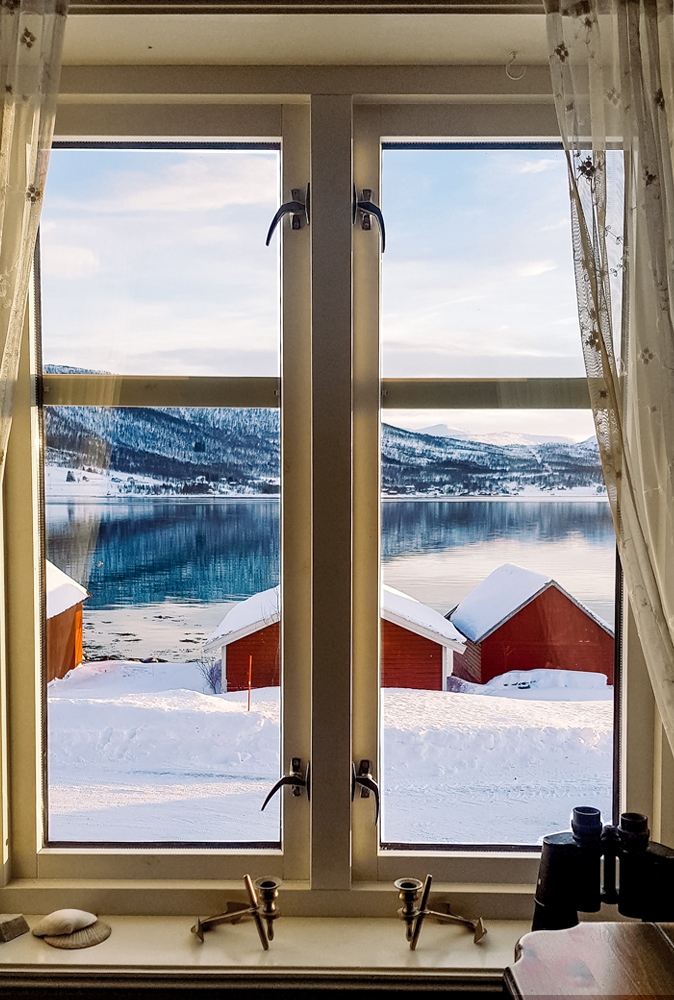 alt=“View of red cabins in snow from the windows in Gibostad town, Northern Norway”