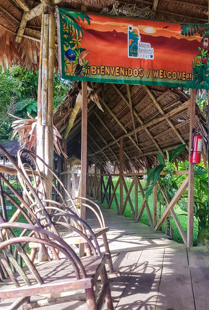 alt=“Amazon Rainforest Lodge entrance walkway with wooden chairs and orange Bienvenidos sign”