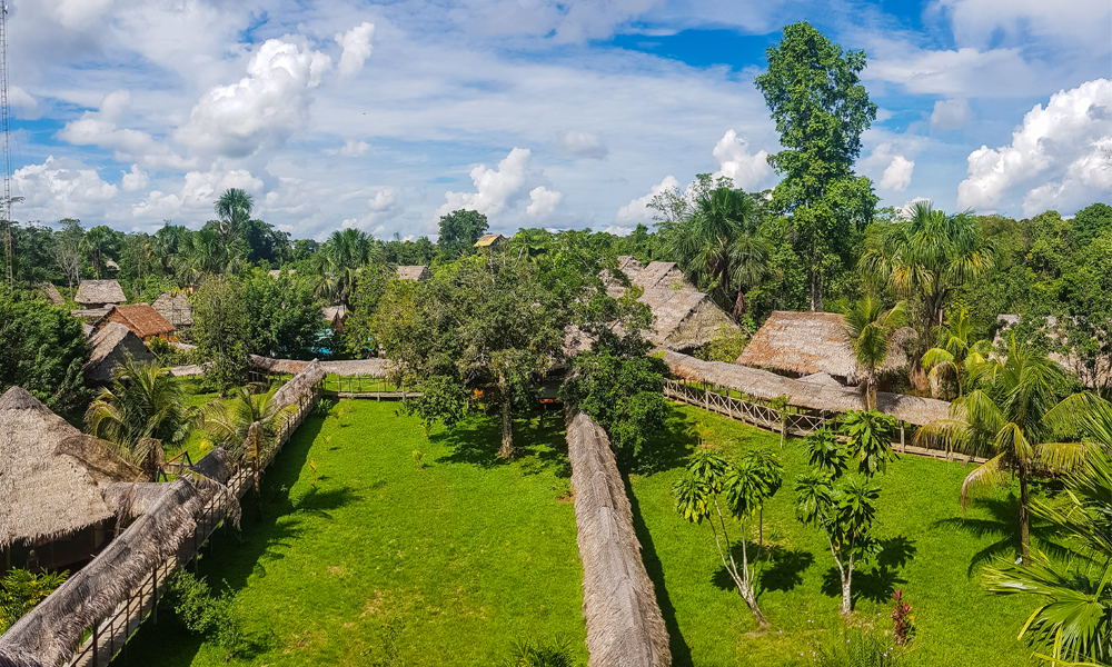alt=“Amazon Rainforest Lodge panoramic view of straw huts and walkway with jungle scenery”