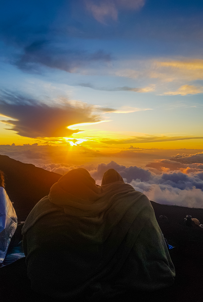 alt="Two girls wrapped up in a blanket watching a beautiful sunset above the clouds on Heleakala Crater in Hawaii"